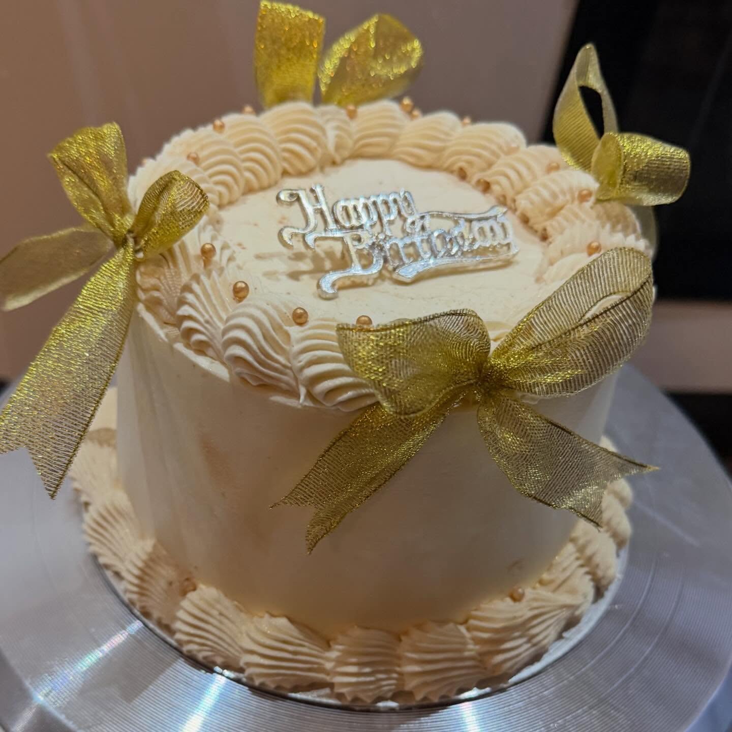 A cream vintage-style cake with gold ribbon bows and a silver Happy Birthday topper